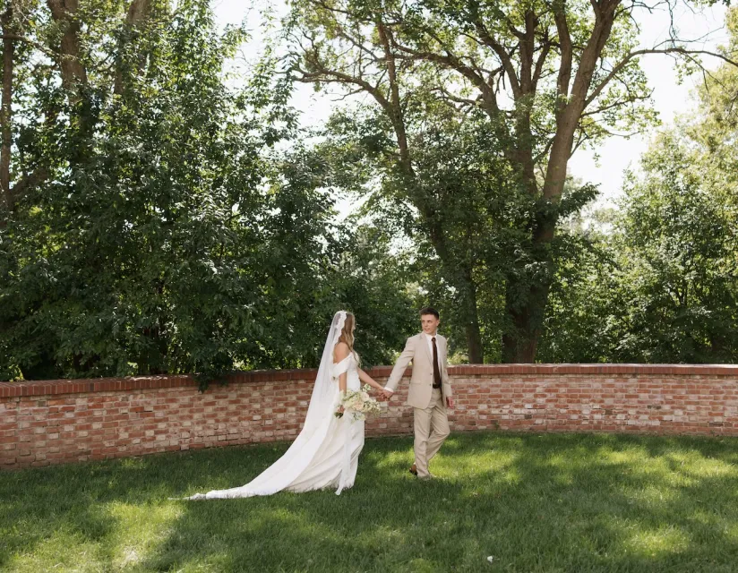 Bride and groom hold hands while walking on grass near a brick wall under tall leafy trees on a sunny day.