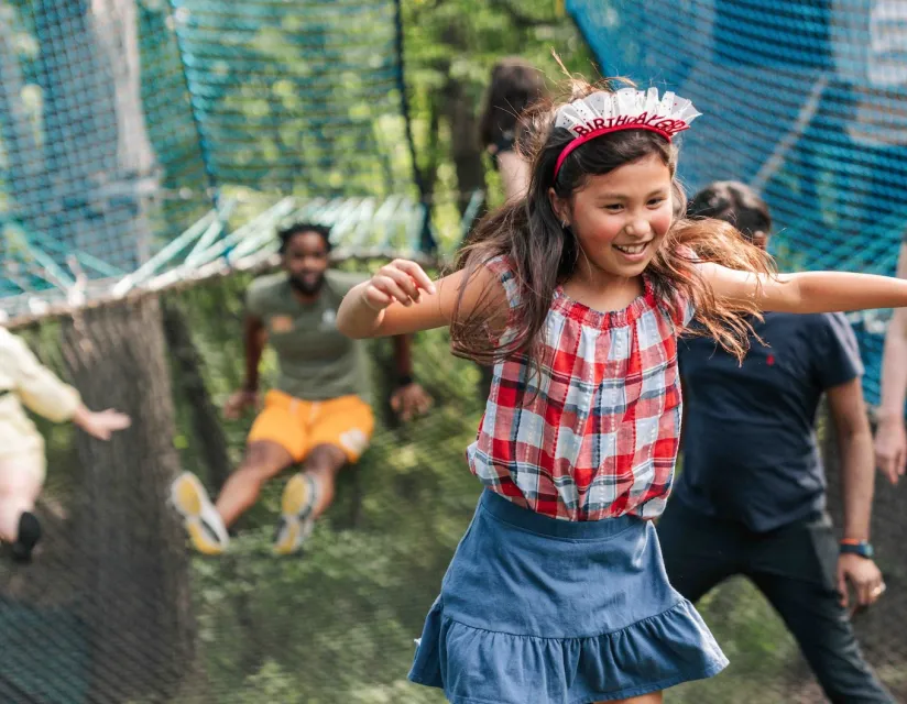 A girl wearing a birthday headband jumps on a trampoline outdoors with several people in the background.