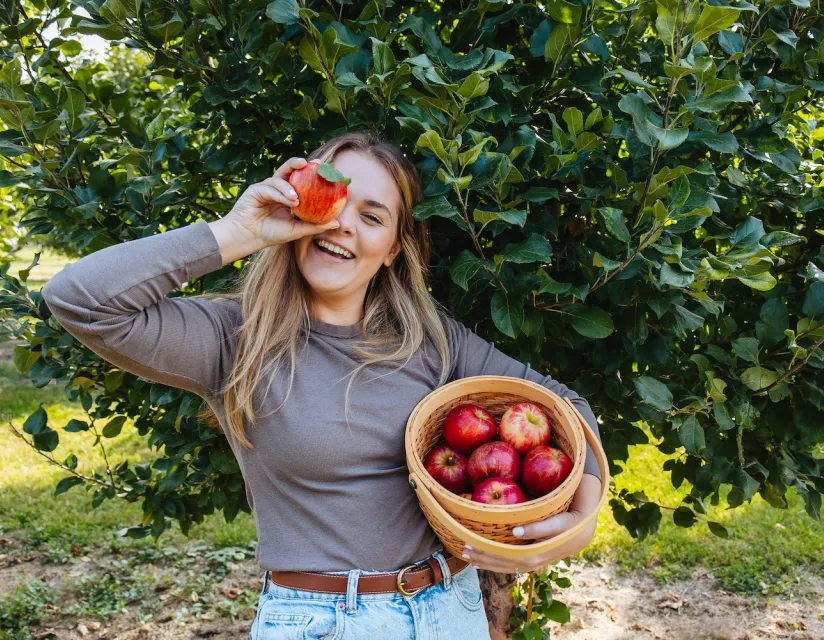 Smiling woman holding a basket of red apples in one arm and playfully holding an apple over one eye in an apple orchard.