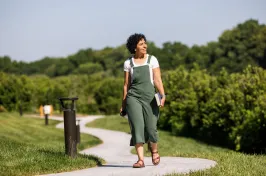 A person in a green jumpsuit walks along a curved pathway in a sunny park, holding a book and a camera. Lush greenery surrounds the path.