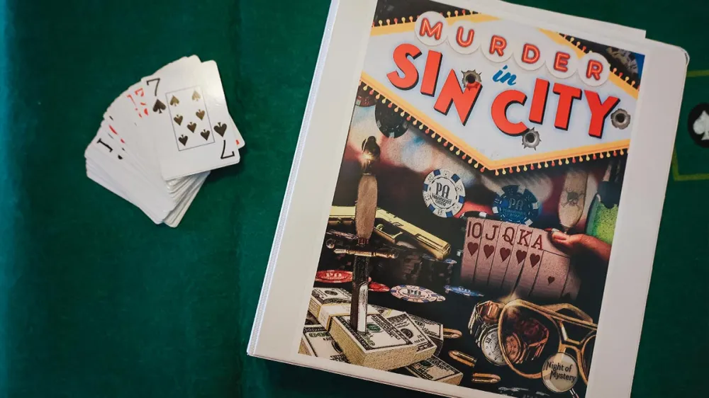 A deck of cards and a binder with 'Murder in Sin City' written on it on a green background.