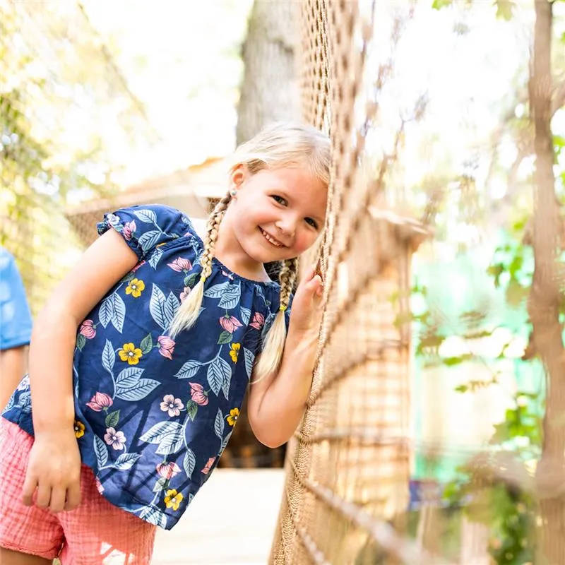 A little girl smiles brightly while standing confidently on a rope bridge, surrounded by greenery.