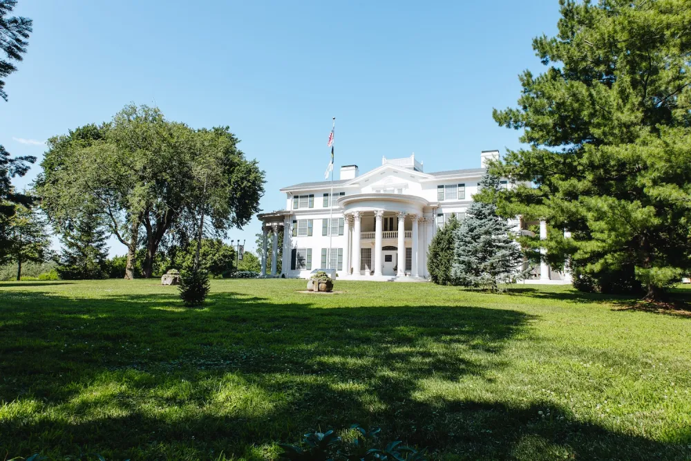 A stately white mansion with columns, surrounded by lush greenery under a clear blue sky, featuring an American flag out front.