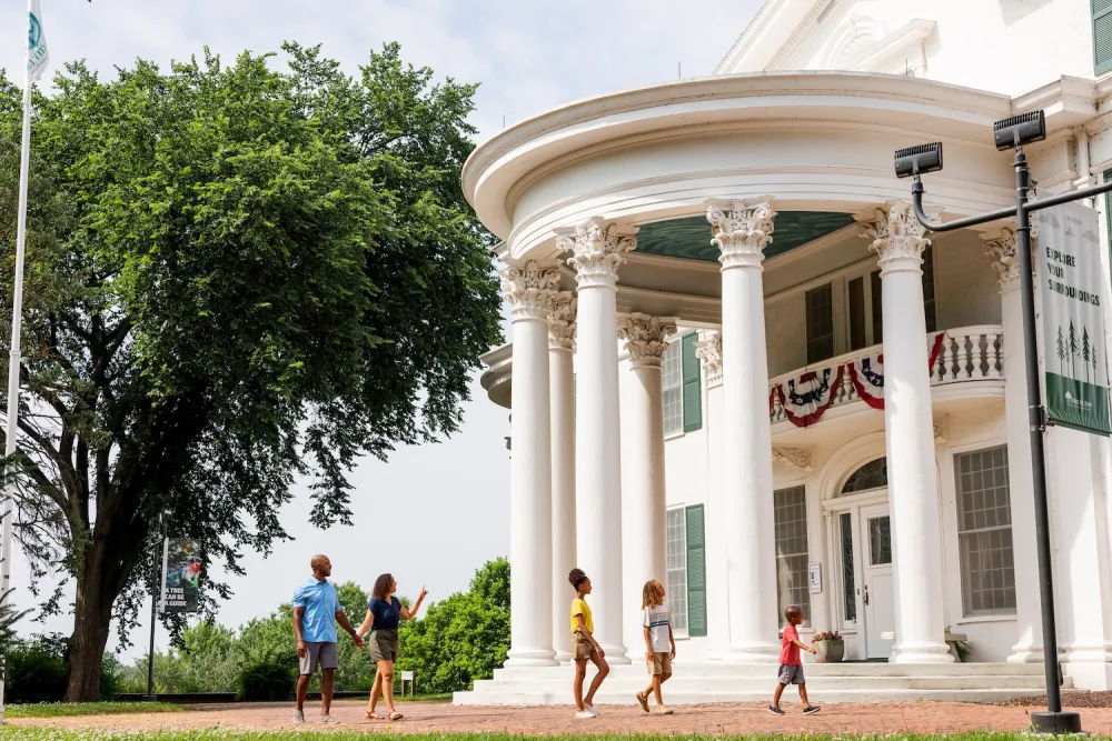 A family of five strolls in front of a grand white building with columns and lush greenery under a partly cloudy sky.