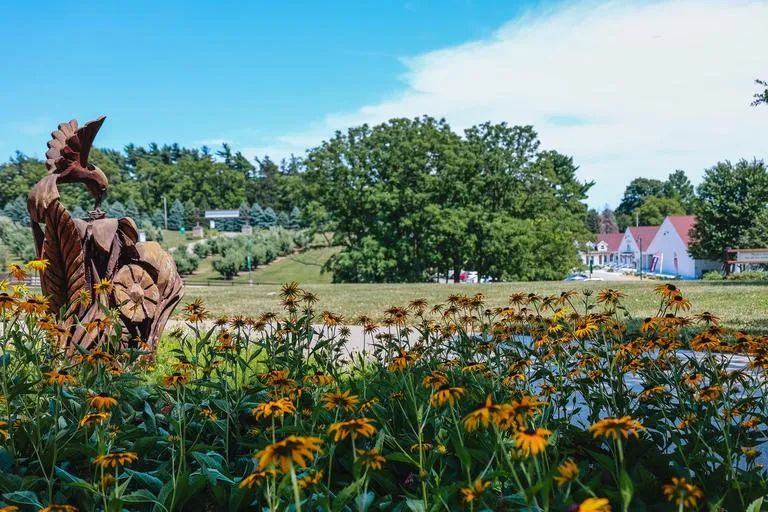 A wooden sculpture of a bird overlooks a vibrant patch of yellow flowers against a scenic hill and blue sky.