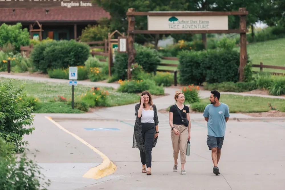 Three people walk along a paved path in a lush park setting, approaching a sign for "Tree Adventure" at Arbor Day Farm.