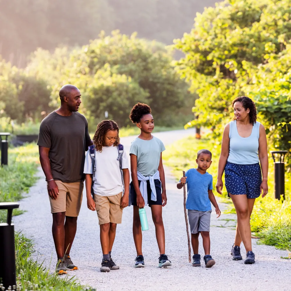 A family of five strolls along a scenic gravel path surrounded by lush greenery, enjoying a sunny day outdoors.