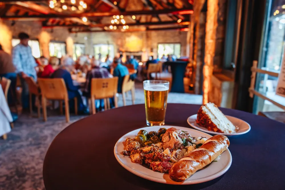 A plate of gourmet food featuring grilled meats, roasted vegetables, a pretzel, and a slice of cake, alongside a glass of beer.