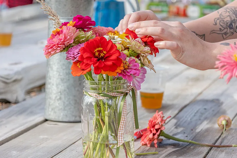 Close up of person arranging bright flowers in a mason jar on a picnic table.