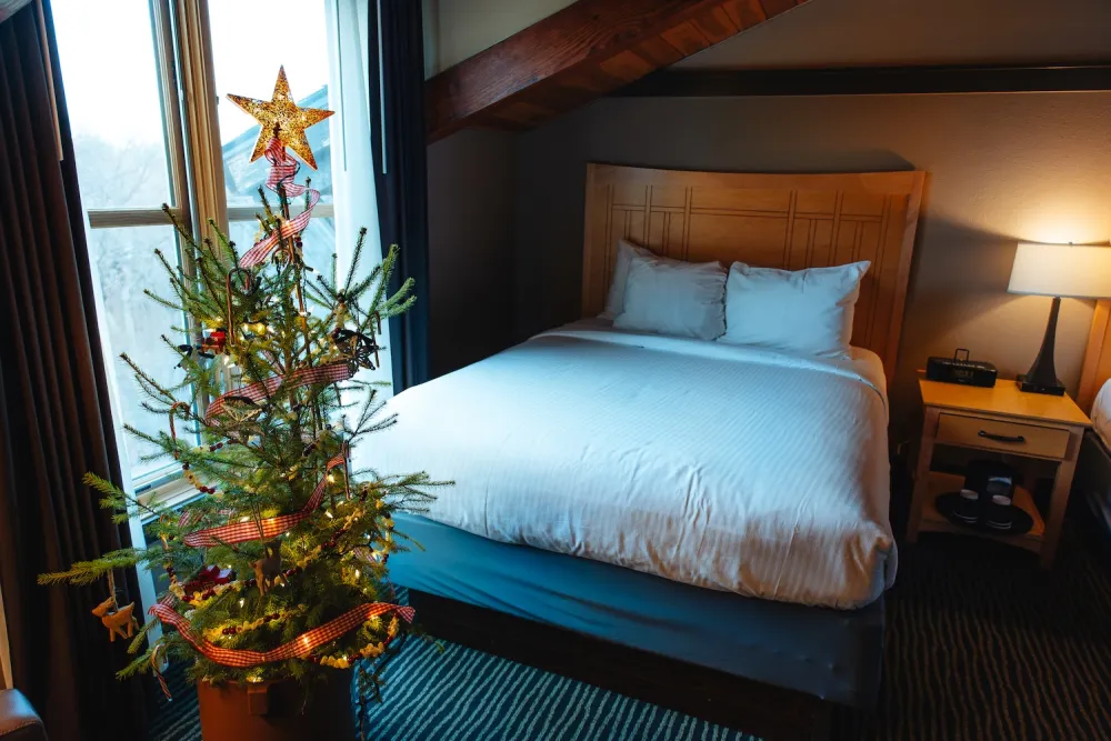 A cozy hotel room featuring a decorated Christmas tree with a star topper, beside a neatly made bed and a bedside lamp.