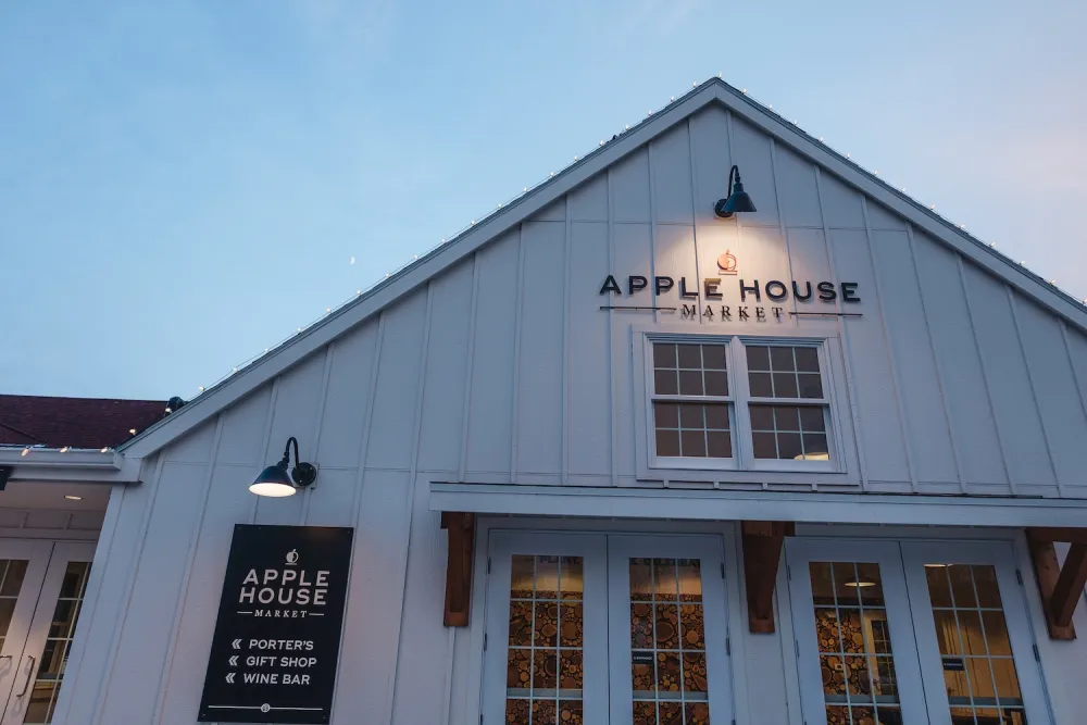 The exterior of Apple House Market, a charming white building with a dark roof, showcasing a sign and warm evening lighting.