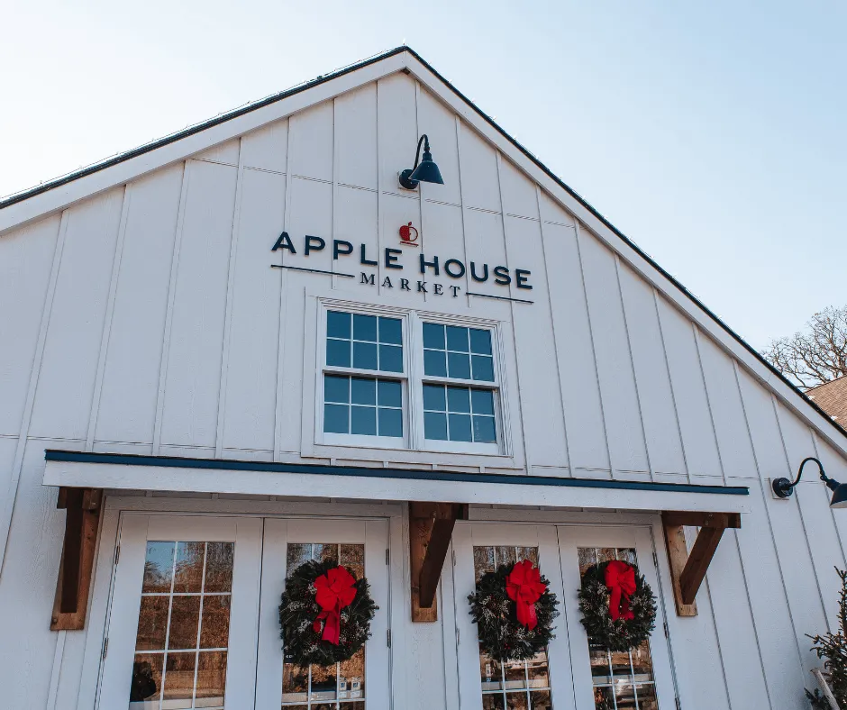 The exterior of Apple House Market features a white facade, two windows, holiday wreaths with red bows on the doors, and a sign above the entrance.