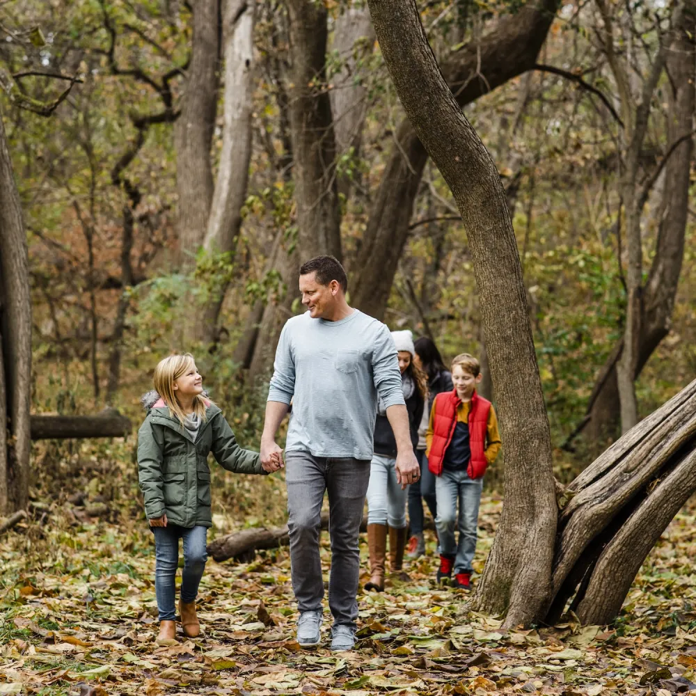 A family walks hand-in-hand through a forested area covered in fallen leaves, surrounded by trees in autumn colors.