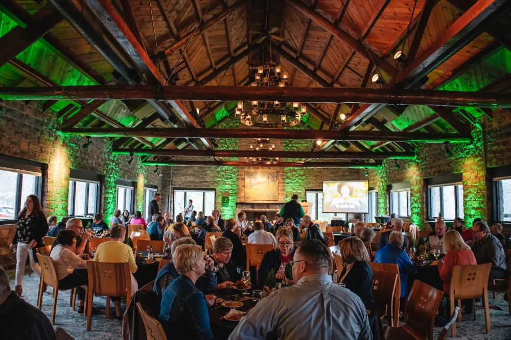 A lively gathering in a rustic, well-lit dining hall with stone walls, wooden beams, and guests enjoying food and drinks.