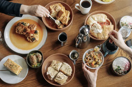 Indoor table filled with brunch items and people eating and serving food in front of them.