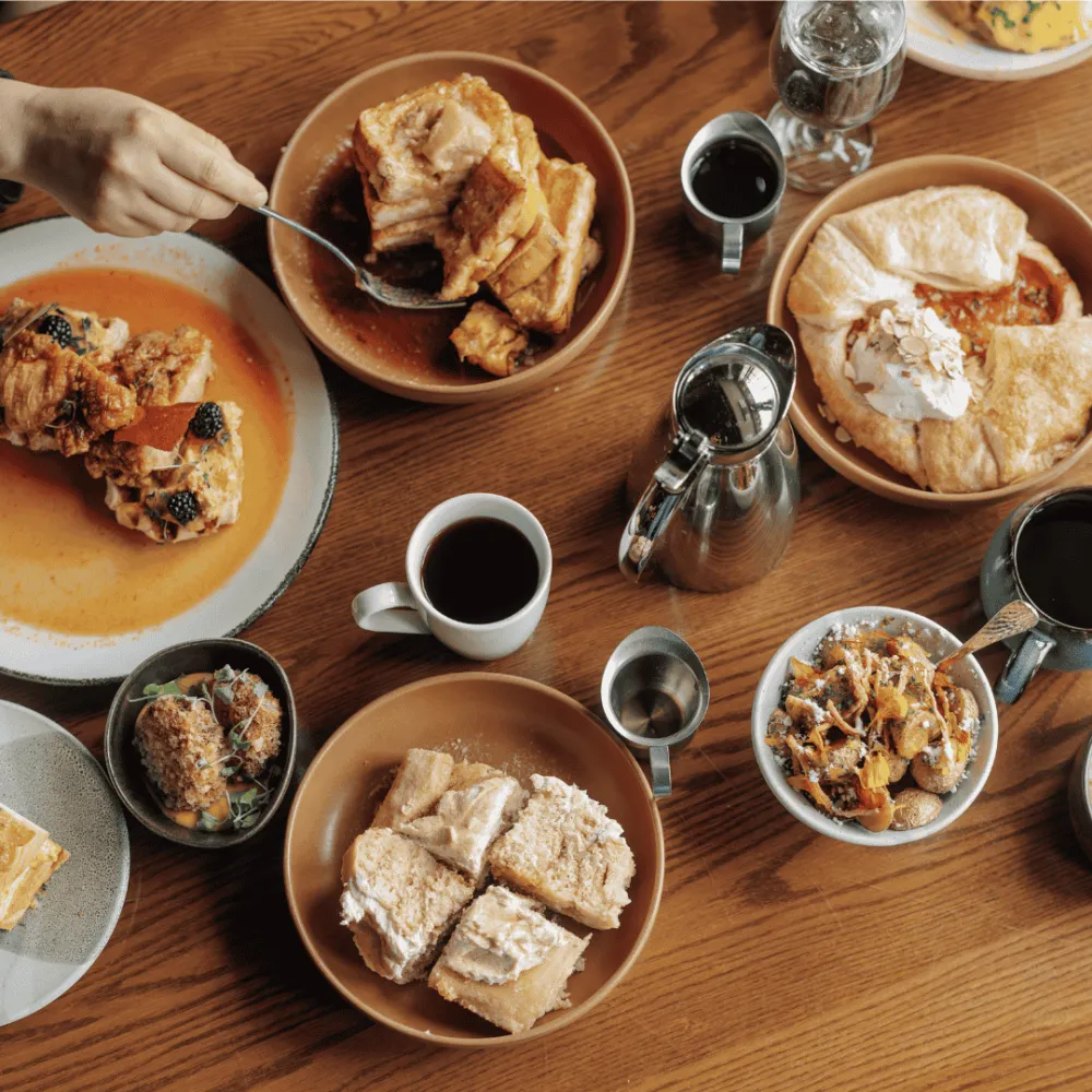 Wooden table with a wide variety of brunch food items and people using forks to eat.