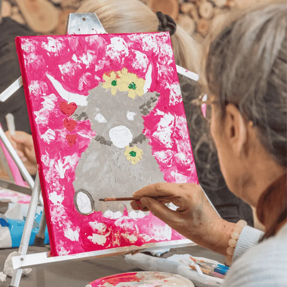 Image of a lady painting in a painting class and is painting a highland coo.