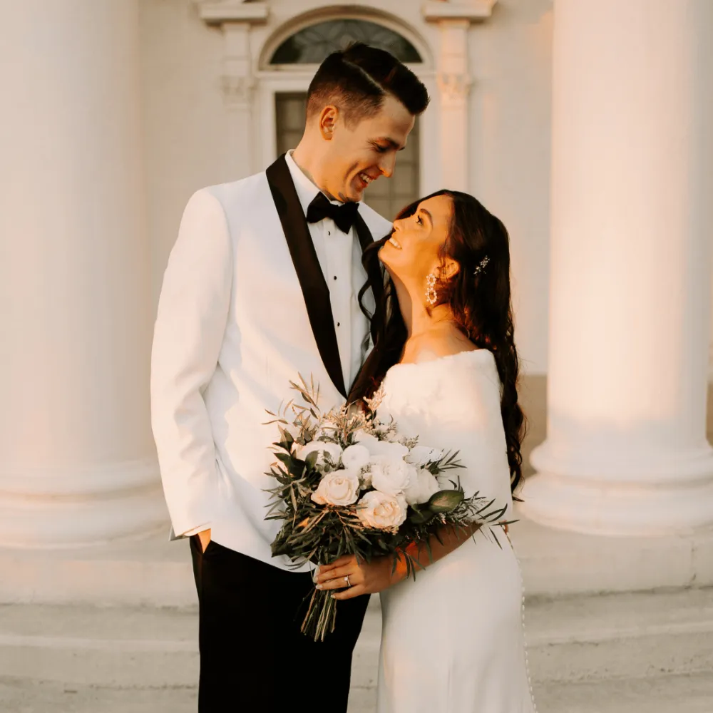 Bride and groom looking at each other in front of a white building with the sun setting.