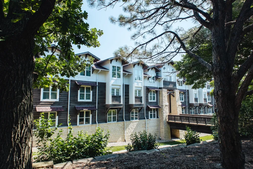 A modern, multi-story building with a mix of stone and wood siding, framed by lush greenery and trees in a sunny setting.