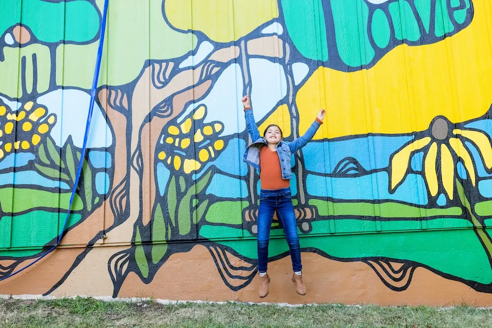 Little girl in jeans, a jean jacket, and red shirt jumping up in the hair with her hands raised in the air against a mural background of virbant yellows, greens, blues, and black outlines. 
