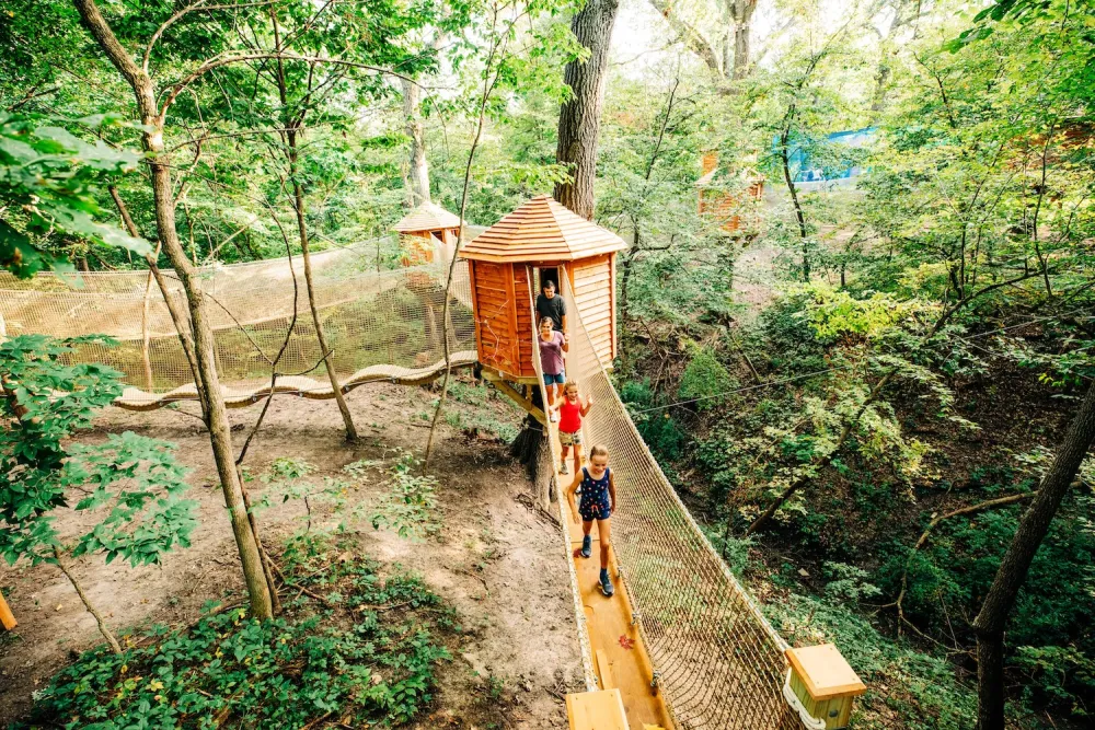Three children and an adult walking on a wooden suspension bridge connected to small treehouse cabins in a forested area.