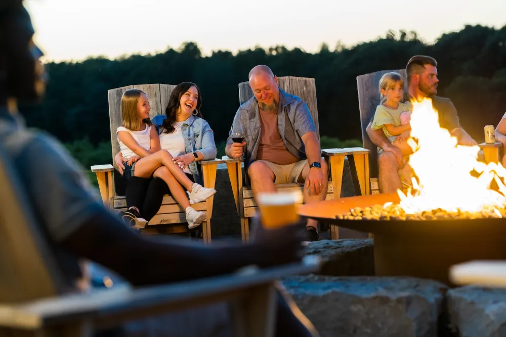 Group of people sitting on wooden chairs around a fire pit outdoors at dusk, holding drinks and enjoying a relaxed evening.