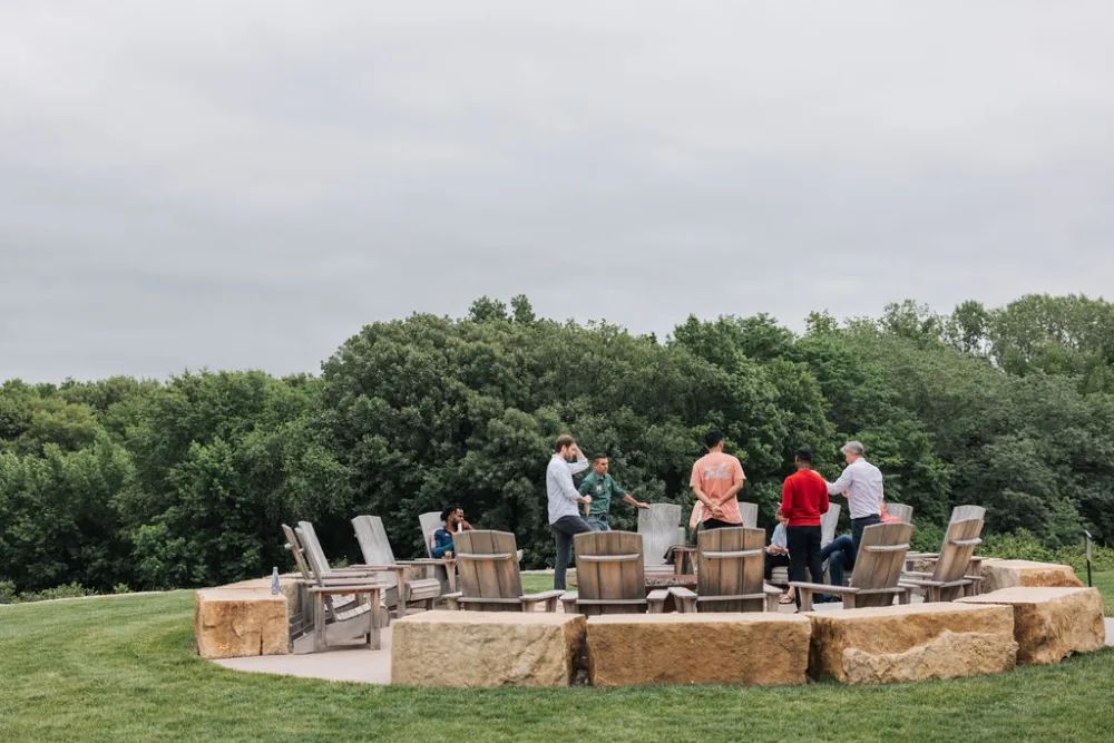 Group of people standing and sitting on wooden chairs in a circular outdoor stone seating area surrounded by greenery on a cloudy day.