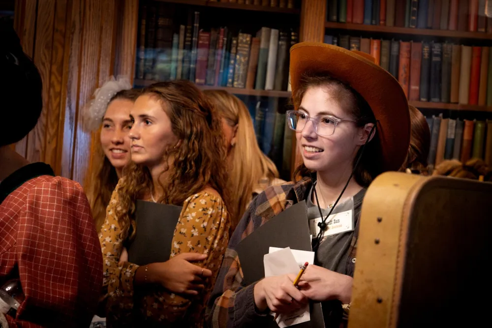 Group of young women in a library, one wearing a cowboy hat and glasses, holding papers and a pencil, engaged in a murder mystery event.