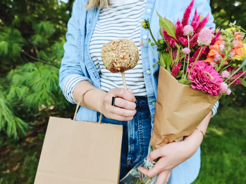A woman holding a caramel apple on a stick and a bouquet of colorful flowers, with a brown paper bag at her side, outdoors.
