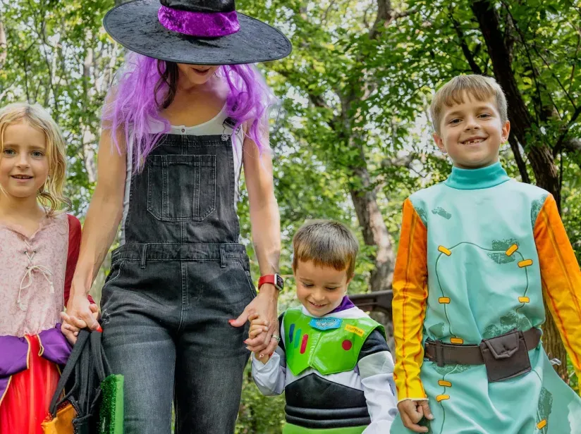 A woman in a black witch hat and purple wig holds hands with three children in colorful costumes, surrounded by greenery.