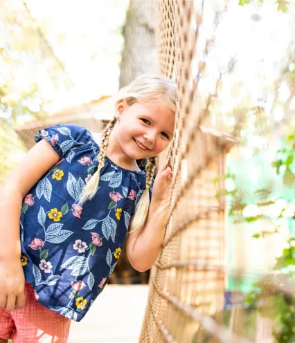 A little girl smiles brightly while standing confidently on a rope bridge, surrounded by greenery.