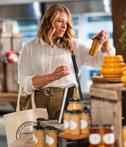 A woman inspects a jar of honey at a market stall filled with various jars and bottles of preserves and honey products.