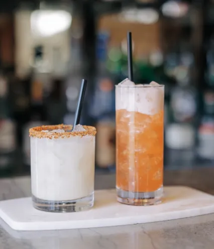 Two cocktails on a white serving board: a creamy, garnished drink and a vibrant orange beverage with ice, set on a marble table.