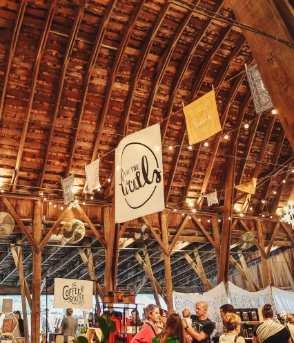 A rustic barn interior filled with vendors, featuring hanging banners, warm lights, and a lively atmosphere.