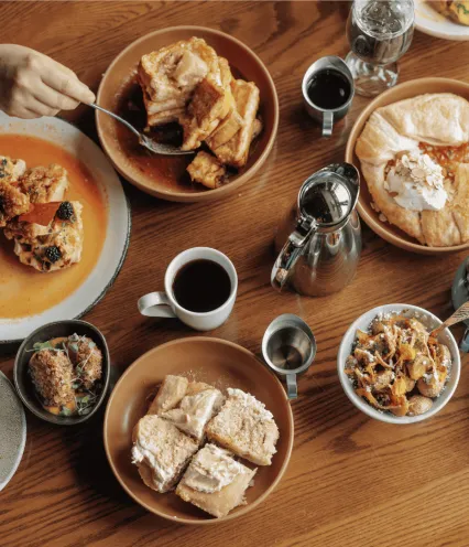 Wooden table with a wide variety of brunch food items and people using forks to eat.