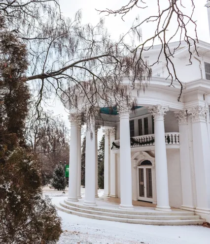 Elegant white building with tall columns and intricate details, surrounded by snow-dusted trees and branches, creating a serene winter scene.