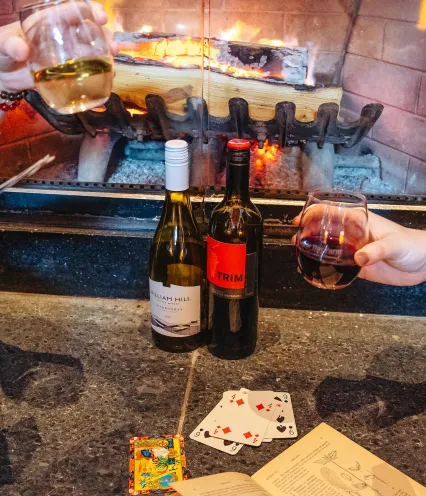 Cozy scene by a fireplace with two wine bottles and glasses, a deck of playing cards, and an open book. Warm, inviting atmosphere.