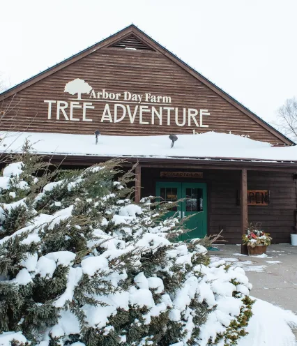 Wooden building labeled "Arbor Day Farm Tree Adventure" in a snowy landscape. Evergreen shrubs with snow in foreground, bare trees in background.