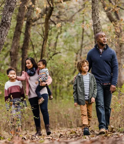 Family of five walking through a forest trail in autumn, surrounded by trees with dry leaves.