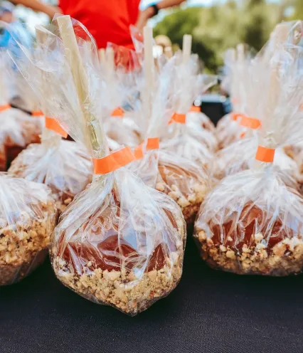 Caramel apples coated with crushed nuts, individually wrapped in plastic with orange ties, displayed on a black tablecloth.