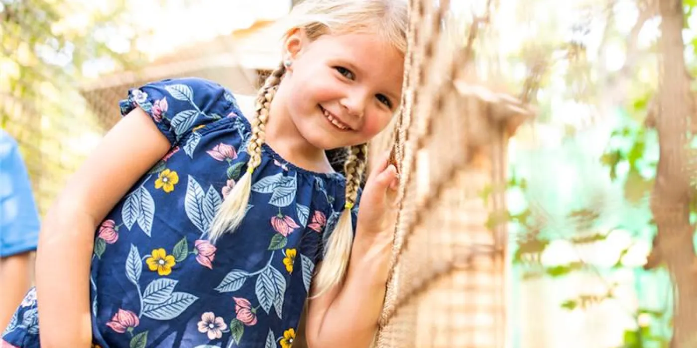 A little girl smiles brightly while standing confidently on a rope bridge, surrounded by greenery.
