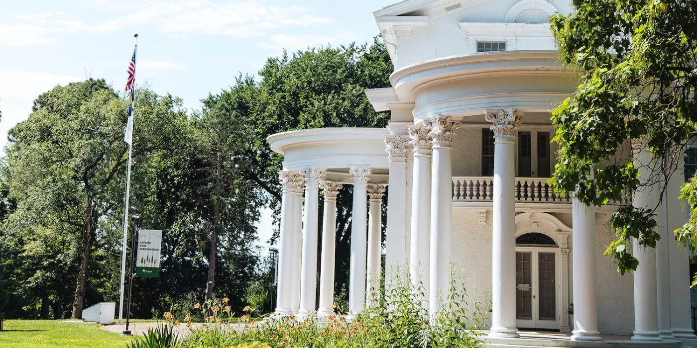 A grand white mansion with Corinthian columns, surrounded by greenery, under a blue sky, showcasing American flag and a sign.