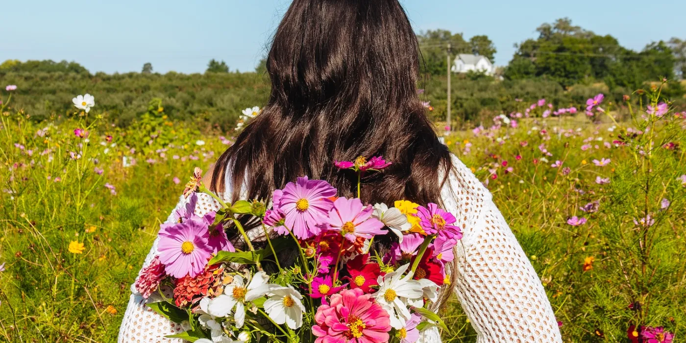 A woman in a white sweater and jeans holds vibrant pink, purple, and white flowers behind her back, standing in a colorful wildflower field under a clear blue sky.