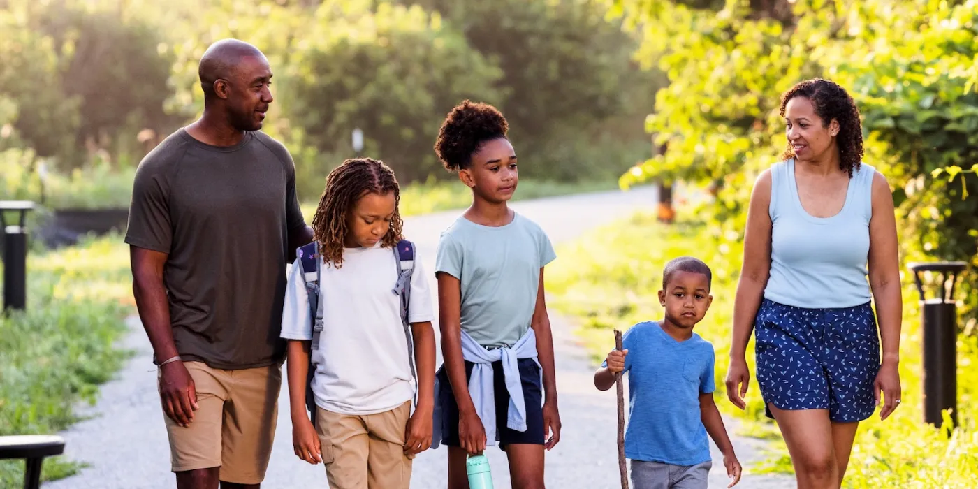 A family of five strolls along a scenic gravel path surrounded by lush greenery, enjoying a sunny day outdoors.