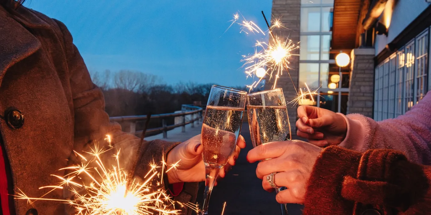 Two people clink champagne glasses, holding sparklers. It's dusk with a festive atmosphere, evoking celebration and joy against a building backdrop.