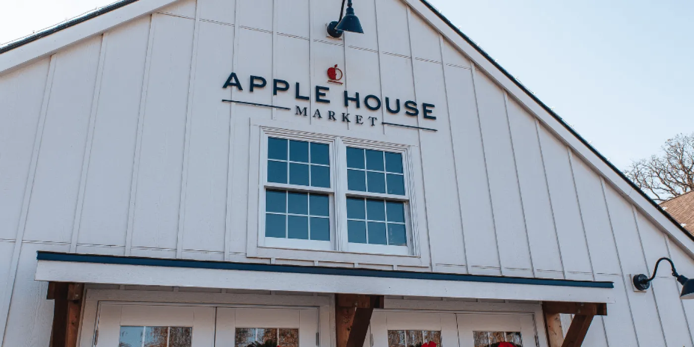 The exterior of Apple House Market features a white facade, two windows, holiday wreaths with red bows on the doors, and a sign above the entrance.