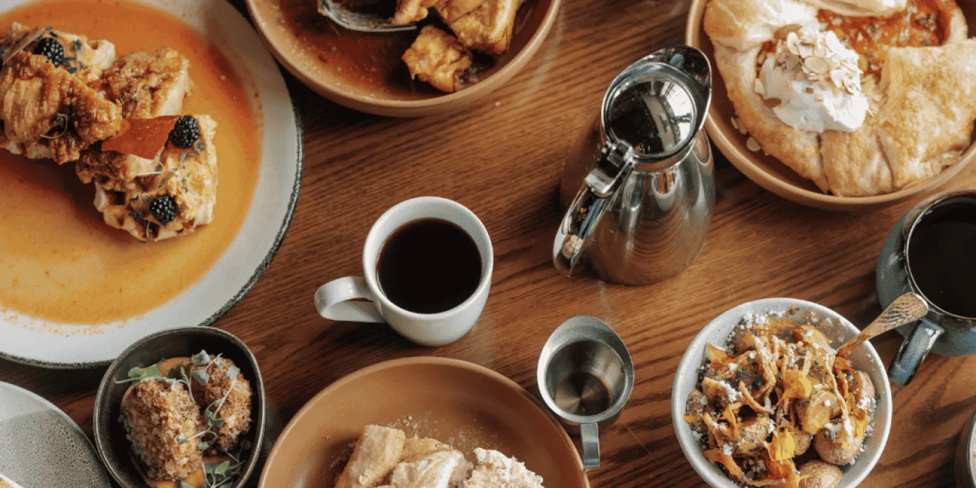 Wooden table with a wide variety of brunch food items and people using forks to eat.