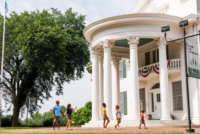 A family of five strolls in front of a grand white building with columns and lush greenery under a partly cloudy sky.
