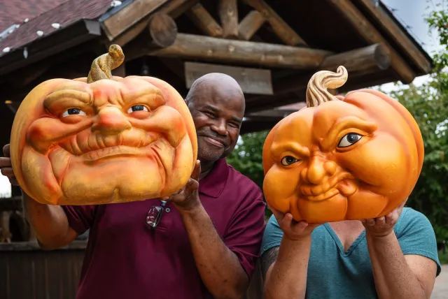 Two people hold uniquely carved, expressive pumpkins in front of a rustic wooden structure surrounded by greenery.