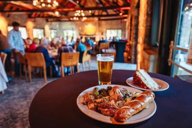 A plate of gourmet food featuring grilled meats, roasted vegetables, a pretzel, and a slice of cake, alongside a glass of beer.